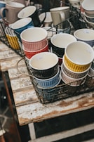 A collection of colorful ceramic mugs stacked inside metal wire baskets, placed on a rustic wooden table. The mugs are in various colors, including red, yellow, blue, and black, each with a ribbed exterior design. Some price tags are visible on the mugs.