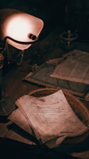 A vintage desk setting featuring a stack of aged documents with handwritten text on rough paper. The scene is dimly lit by an old-fashioned lamp that casts a warm glow across the papers and a woven basket upon which they rest. Dust and signs of age suggest an antiquated or neglected atmosphere.
