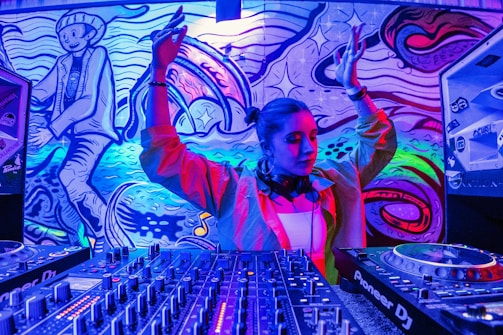 A DJ standing at a set of mixing equipment with a colorful, vibrant mural in the background. The mural features abstract designs and a cartoonish character. The DJ has headphones around their neck and their arms raised, possibly in mid-performance.