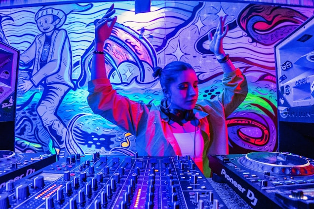 A DJ standing at a set of mixing equipment with a colorful, vibrant mural in the background. The mural features abstract designs and a cartoonish character. The DJ has headphones around their neck and their arms raised, possibly in mid-performance.