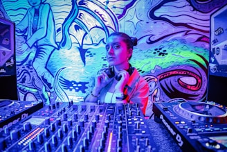 A woman is surrounded by DJ equipment, including a mixer and turntables, against a vibrant, graffiti-covered wall with abstract designs. She wears headphones around her neck and looks confidently at the camera.