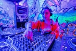 A DJ wearing headphones operates a sound mixer in a vibrant, graffiti-decorated environment. The scene is illuminated with neon lights, creating a vivid blue and pink hue. The graffiti on the walls features artistic and musical imagery.