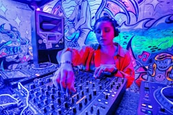 A DJ wearing headphones operates a sound mixer in a vibrant, graffiti-decorated environment. The scene is illuminated with neon lights, creating a vivid blue and pink hue. The graffiti on the walls features artistic and musical imagery.