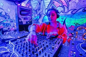 A DJ wearing headphones operates a sound mixer in a vibrant, graffiti-decorated environment. The scene is illuminated with neon lights, creating a vivid blue and pink hue. The graffiti on the walls features artistic and musical imagery.