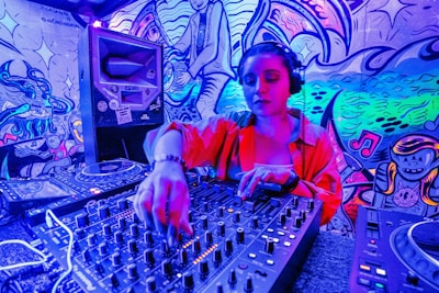 A DJ wearing headphones operates a sound mixer in a vibrant, graffiti-decorated environment. The scene is illuminated with neon lights, creating a vivid blue and pink hue. The graffiti on the walls features artistic and musical imagery.
