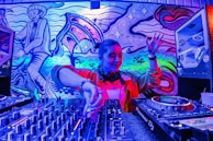 A DJ is performing, skillfully mixing tracks on a large set of DJ equipment. The background features vibrant, colorful street art, including abstract designs and a stylized character. The scene is illuminated by intense blue, purple, and pink lighting.