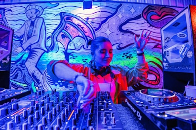 A DJ is performing, skillfully mixing tracks on a large set of DJ equipment. The background features vibrant, colorful street art, including abstract designs and a stylized character. The scene is illuminated by intense blue, purple, and pink lighting.