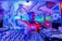 A DJ is performing, skillfully mixing tracks on a large set of DJ equipment. The background features vibrant, colorful street art, including abstract designs and a stylized character. The scene is illuminated by intense blue, purple, and pink lighting.