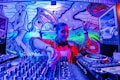 A DJ is performing, skillfully mixing tracks on a large set of DJ equipment. The background features vibrant, colorful street art, including abstract designs and a stylized character. The scene is illuminated by intense blue, purple, and pink lighting.