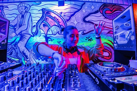 A DJ is performing, skillfully mixing tracks on a large set of DJ equipment. The background features vibrant, colorful street art, including abstract designs and a stylized character. The scene is illuminated by intense blue, purple, and pink lighting.