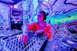 A person is DJing in a vibrant room filled with colorful graffiti art on the walls. The individual is wearing headphones and operating a sound mixer. The background features playful cartoonish characters and musical notes in a dynamic, swirling pattern.