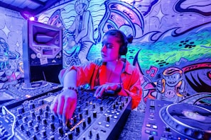 A person is DJing in a vibrant room filled with colorful graffiti art on the walls. The individual is wearing headphones and operating a sound mixer. The background features playful cartoonish characters and musical notes in a dynamic, swirling pattern.