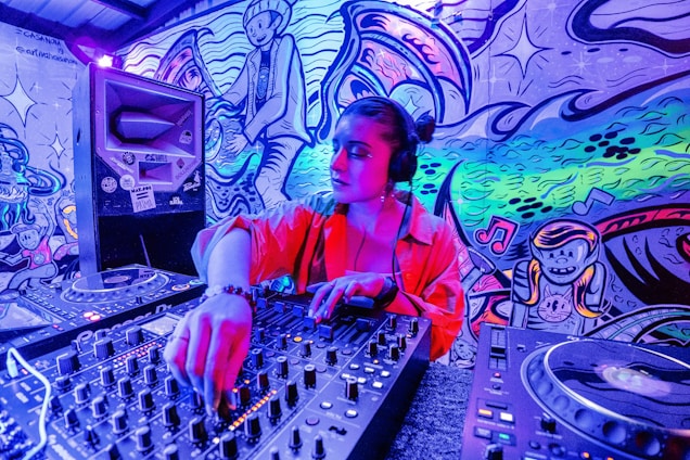 A person is DJing in a vibrant room filled with colorful graffiti art on the walls. The individual is wearing headphones and operating a sound mixer. The background features playful cartoonish characters and musical notes in a dynamic, swirling pattern.