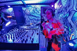 A DJ booth is set up in an urban setting with a colorful, graffiti-covered wall as a backdrop. The DJ is energetically performing, wearing headphones and gesturing with their hand. The scene is illuminated by vibrant blue and red lighting, creating an electrifying atmosphere. In the foreground, a large mixer and sound equipment are visible.
