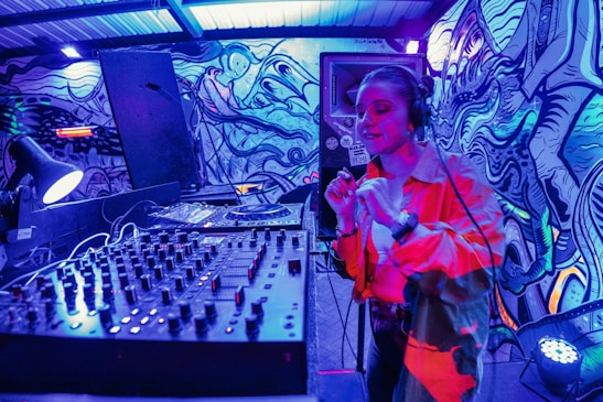 A DJ booth is set up in an urban setting with a colorful, graffiti-covered wall as a backdrop. The DJ is energetically performing, wearing headphones and gesturing with their hand. The scene is illuminated by vibrant blue and red lighting, creating an electrifying atmosphere. In the foreground, a large mixer and sound equipment are visible.