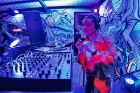A DJ booth is set up in an urban setting with a colorful, graffiti-covered wall as a backdrop. The DJ is energetically performing, wearing headphones and gesturing with their hand. The scene is illuminated by vibrant blue and red lighting, creating an electrifying atmosphere. In the foreground, a large mixer and sound equipment are visible.