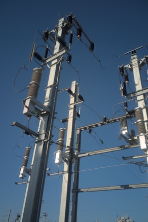 Tall electric power transmission towers with various wires and insulators are seen against a clear blue sky. The towers are composed of metallic structures with cylindrical insulators and other electrical components attached at different heights.