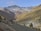 A picturesque mountain landscape features snow-capped peaks in the background with a verdant valley below. Autumn-colored trees blanket the slopes, and a small village is nestled in the valley, adding charm to the serene scene.