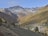 A picturesque mountain landscape features snow-capped peaks in the background with a verdant valley below. Autumn-colored trees blanket the slopes, and a small village is nestled in the valley, adding charm to the serene scene.