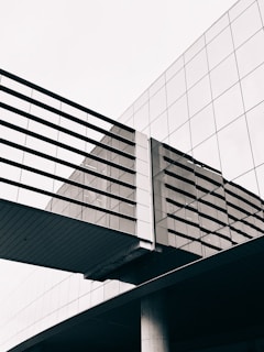 An architectural structure features a modern glass building with reflective windows and a suspended pedestrian bridge. The geometric lines and angles create a sleek and contemporary appearance, emphasizing symmetry and clean design.