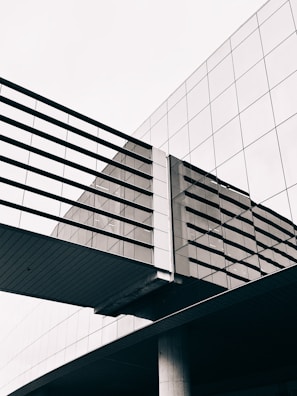 An architectural structure features a modern glass building with reflective windows and a suspended pedestrian bridge. The geometric lines and angles create a sleek and contemporary appearance, emphasizing symmetry and clean design.