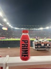 A red bottle labeled 'PRIME Hydration 500 mL Tropical Punch' is positioned prominently on a white railing. In the background, a well-lit sports stadium is visible, with a large crowd and field activities occurring under the night sky.
