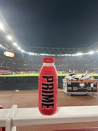 A red bottle labeled 'PRIME Hydration 500 mL Tropical Punch' is positioned prominently on a white railing. In the background, a well-lit sports stadium is visible, with a large crowd and field activities occurring under the night sky.