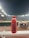 A red bottle labeled 'PRIME Hydration 500 mL Tropical Punch' is positioned prominently on a white railing. In the background, a well-lit sports stadium is visible, with a large crowd and field activities occurring under the night sky.