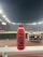 A red bottle labeled 'PRIME Hydration 500 mL Tropical Punch' is positioned prominently on a white railing. In the background, a well-lit sports stadium is visible, with a large crowd and field activities occurring under the night sky.