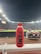 A red bottle labeled 'PRIME Hydration 500 mL Tropical Punch' is positioned prominently on a white railing. In the background, a well-lit sports stadium is visible, with a large crowd and field activities occurring under the night sky.
