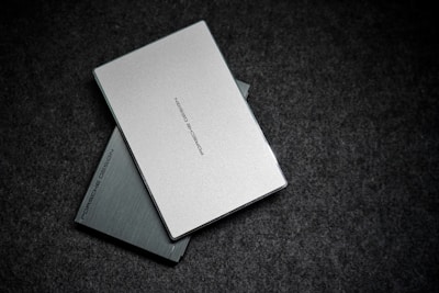 Two sleek, minimalist, rectangular objects with a metallic finish arranged on a textured dark surface. The top object is a light silver color, overlaid on a darker silver or grey one, both featuring the brand name 'Porsche Design' printed in subtle, elegant lettering.