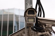 A collection of surveillance cameras mounted on a pole, with various wires and attachments visible. The cameras are positioned near a modern glass building, partially visible in the background.