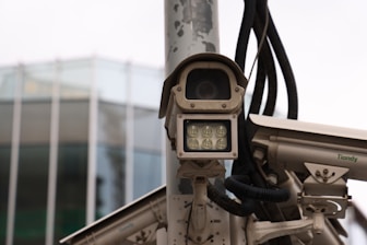 A collection of surveillance cameras mounted on a pole, with various wires and attachments visible. The cameras are positioned near a modern glass building, partially visible in the background.