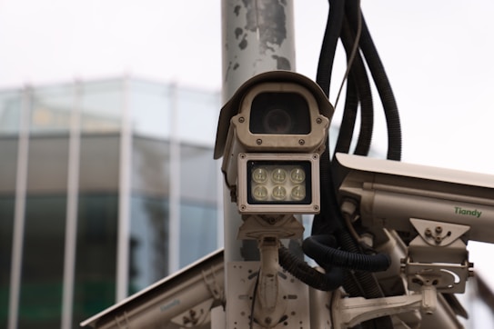 A collection of surveillance cameras mounted on a pole, with various wires and attachments visible. The cameras are positioned near a modern glass building, partially visible in the background.