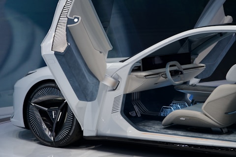 A modern, futuristic car interior featuring sleek design elements. The open door reveals a minimalistic steering wheel and a large, horizontal display. Upholstered seating and a detailed dashboard enhance the advanced technological look. The car has an elegant white exterior with unique alloy wheel design.