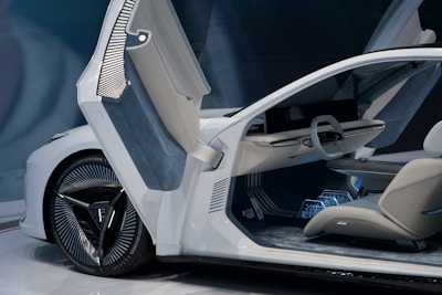 A modern, futuristic car interior featuring sleek design elements. The open door reveals a minimalistic steering wheel and a large, horizontal display. Upholstered seating and a detailed dashboard enhance the advanced technological look. The car has an elegant white exterior with unique alloy wheel design.