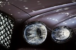 Close-up of a luxury car's front, focusing on the intricate design of the headlights and the chrome grille. The surface reflects ambient lights, creating a sleek and polished appearance.