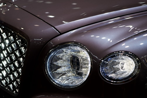 Close-up of a luxury car's front, focusing on the intricate design of the headlights and the chrome grille. The surface reflects ambient lights, creating a sleek and polished appearance.
