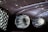Close-up of a luxury car's front, focusing on the intricate design of the headlights and the chrome grille. The surface reflects ambient lights, creating a sleek and polished appearance.