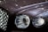 Close-up of a luxury car's front, focusing on the intricate design of the headlights and the chrome grille. The surface reflects ambient lights, creating a sleek and polished appearance.