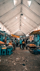 A busy indoor market with people actively shopping at various stalls. The high ceiling with triangular skylights allows natural light to filter through, and the aisles are lined with baskets of fruits and vegetables. Vendors are attending to customers amidst a rustic and slightly chaotic atmosphere.