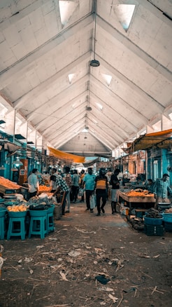 A busy indoor market with people actively shopping at various stalls. The high ceiling with triangular skylights allows natural light to filter through, and the aisles are lined with baskets of fruits and vegetables. Vendors are attending to customers amidst a rustic and slightly chaotic atmosphere.