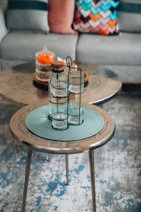 A detailed image of a cozy living room setting with two intricately designed coffee tables. The foreground table features a glass holder with two cylindrical glasses on a textured blue placemat. The background table has a decorative tray topped with a glass container filled with orange flowers and another with a cork top. A stylish sofa with multiple cushions, including a multi-colored chevron-patterned pillow, adds a touch of warmth and color.