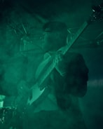 A person is playing a bass guitar on stage, surrounded by green lighting and fog, creating a moody atmosphere. The lighting highlights the details of the guitar and casts shadows on the person, who appears to be focused on the performance.