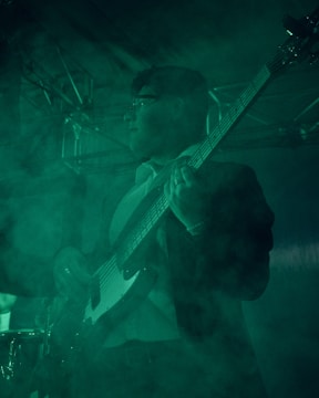 A person is playing a bass guitar on stage, surrounded by green lighting and fog, creating a moody atmosphere. The lighting highlights the details of the guitar and casts shadows on the person, who appears to be focused on the performance.