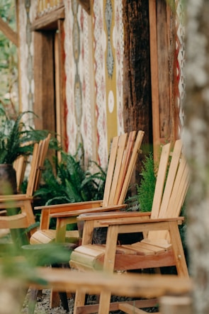 Wooden chairs are arranged in a rustic outdoor setting, flanked by lush greenery and ferns. The backdrop features a wall with colorful, geometric patterns. The overall ambience suggests a tranquil, natural environment.
