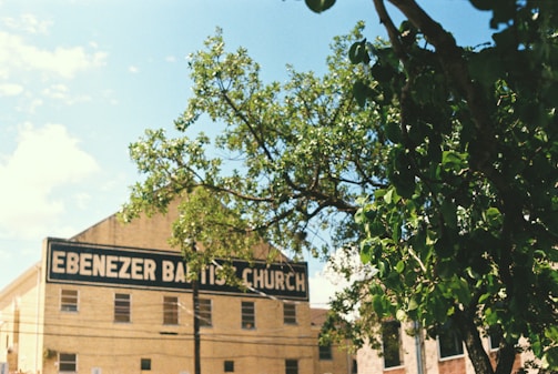 A light-colored building with a sign that reads 'Ebenezer Baptist Church' is partially obscured by a leafy tree. The structure is set against a clear blue sky, evoking a serene and sunny day. Multiple small rectangular windows can be seen on the building's facade.