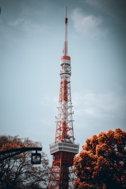 A tall red and white broadcast tower stands prominently against a cloudy sky, surrounded by autumnal trees with orange foliage.