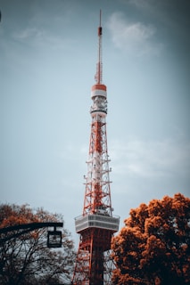 A tall red and white broadcast tower stands prominently against a cloudy sky, surrounded by autumnal trees with orange foliage.