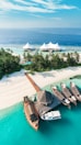 A tropical island features pristine white sandy beaches, lush green palm trees, and turquoise blue waters. A group of traditional wooden boats and a small yacht are docked at a wooden pier. In the background, a luxury resort with white tent-like structures is visible on the water.